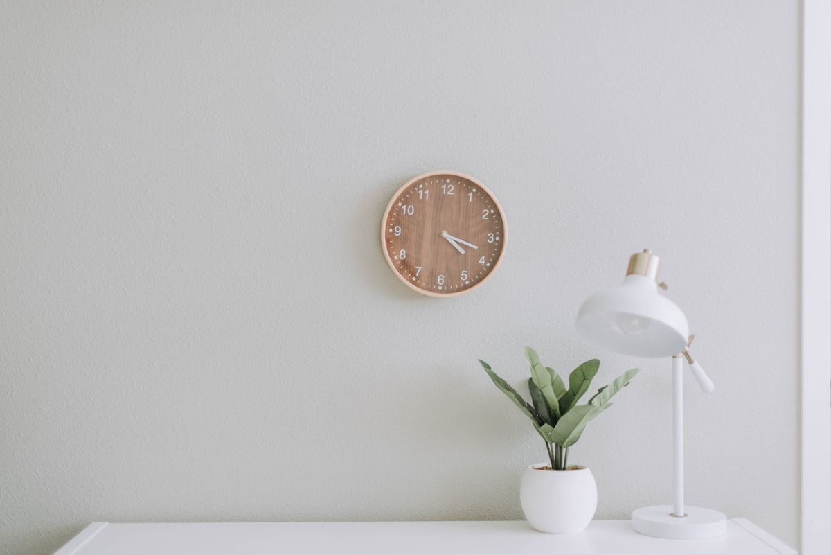 A white desk corner with a leafy plant, desk lamp, and wooden wall clock.